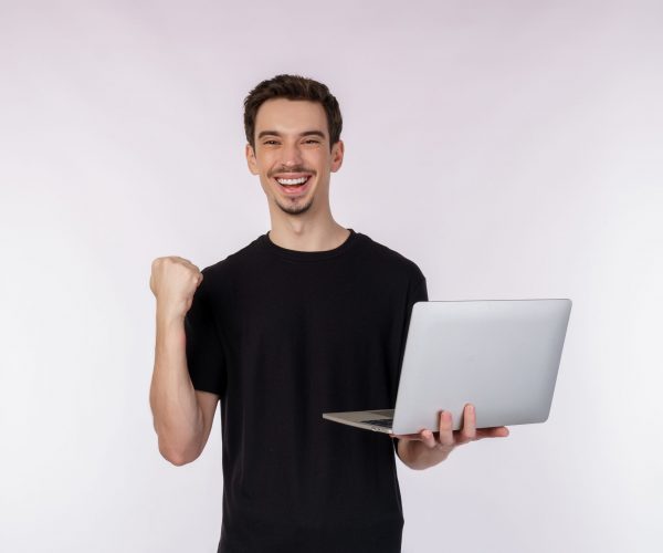 Portrait of young handsome smiling man holding laptop in hands, typing and browsing web pages while doing a winning closed fist gesture isolated on white background. Technology and business concept.